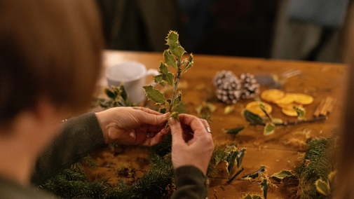 Close-up of hands arranging holly sprigs into a natural decoration at a wooden table.